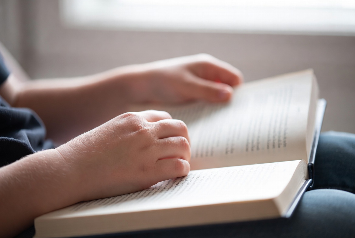 A close-up of a child's hands holding an open paperback book in soft window light.
